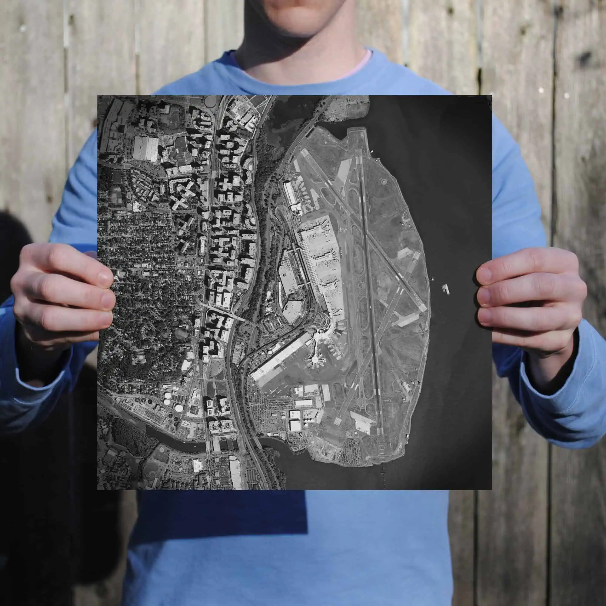 Detailed aerial black and white map of an urban area with roads, buildings, and a river, held by a person in a blue jacket standing outdoors against a wooden fence.