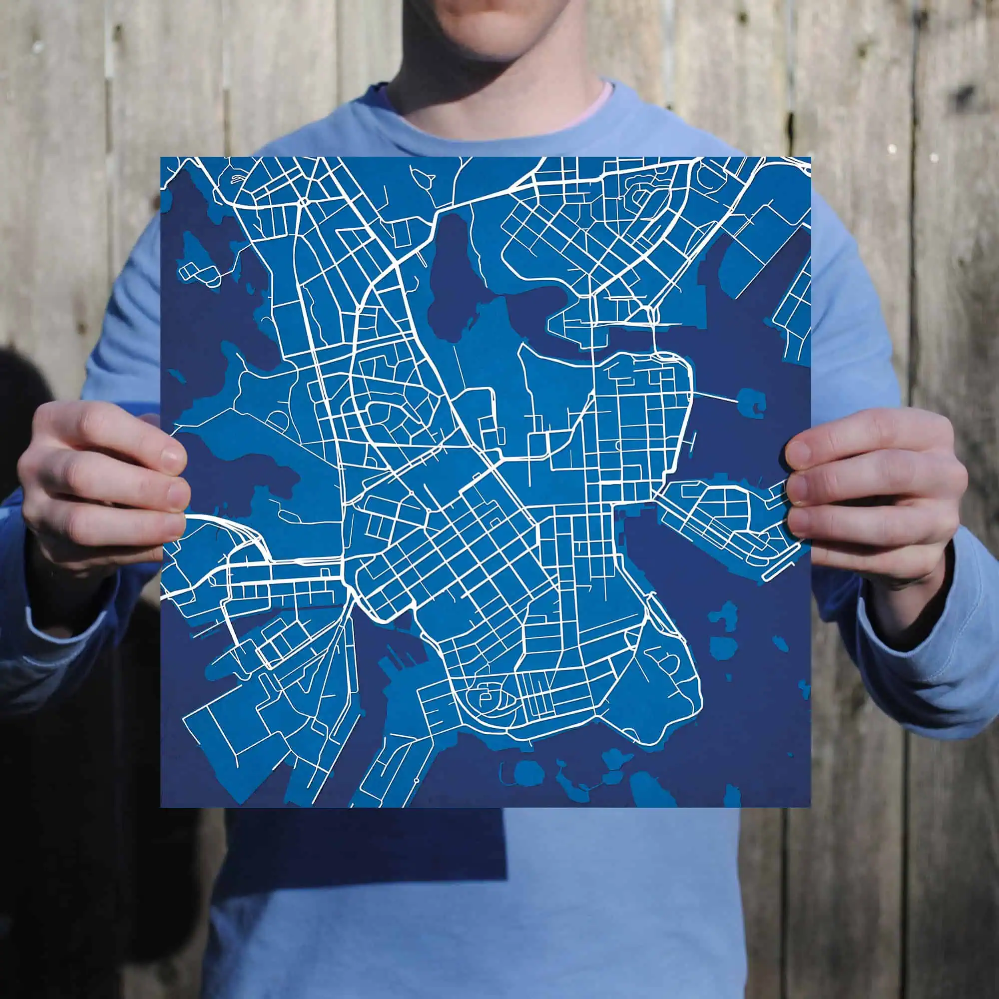 Blue city street map poster held by a person in front of a wooden background, ideal for travel, navigation, and geography enthusiasts.