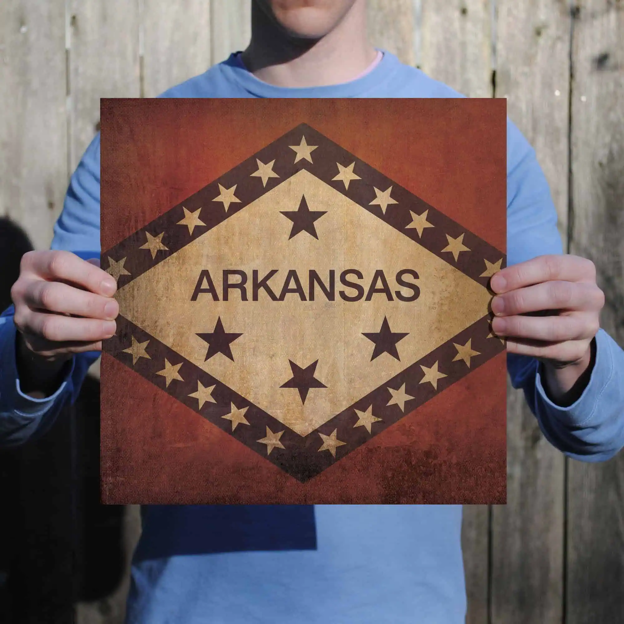 Vintage Arkansas state flag with stars and rustic design, held by a person in front of a wooden fence. Ideal for travel, tourism, or regional pride themes related to Arkansas.