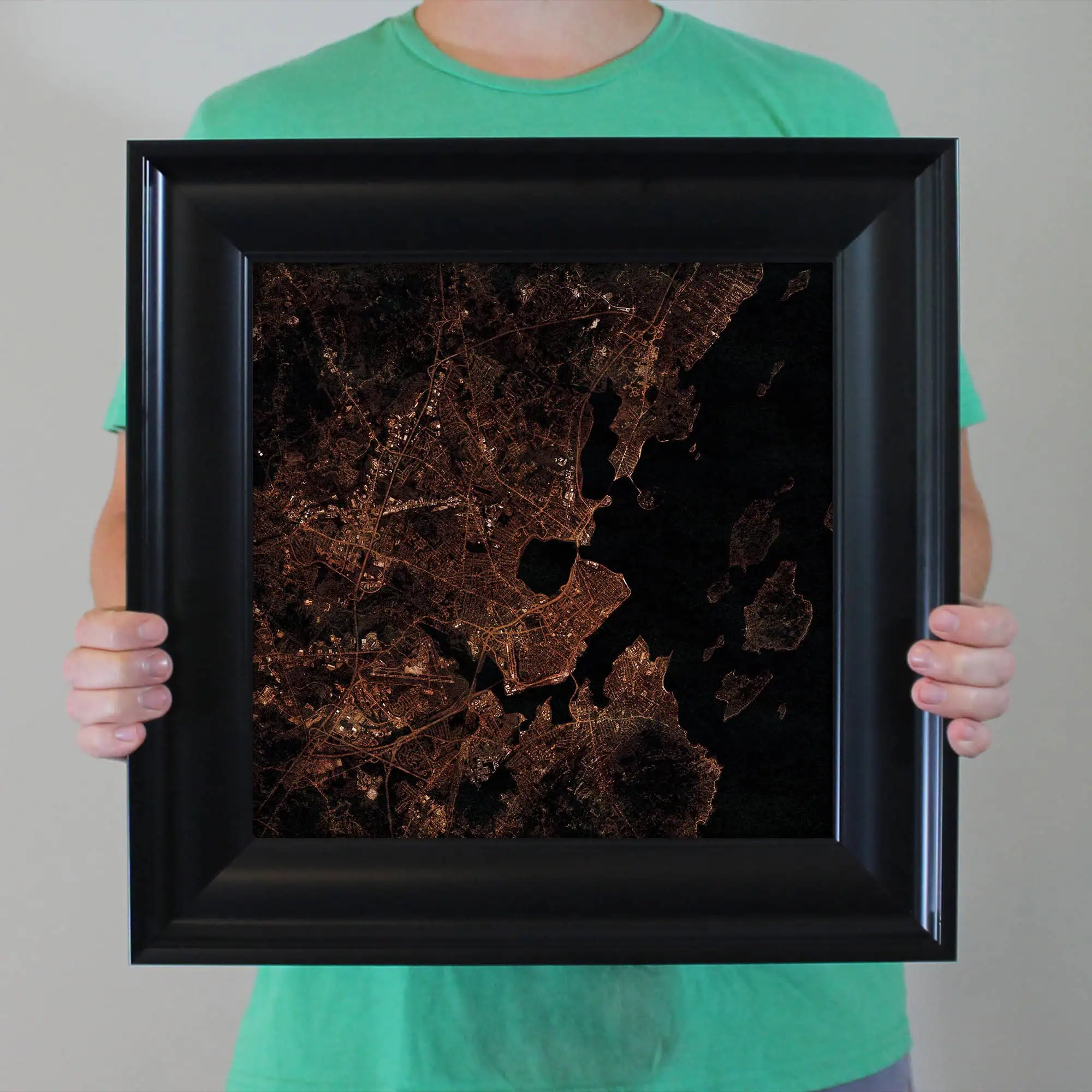 Dark themed city map artwork with illuminated street details, held in a black frame by a person wearing a green t-shirt. This modern art piece showcases detailed urban mapping perfect for wall decor.