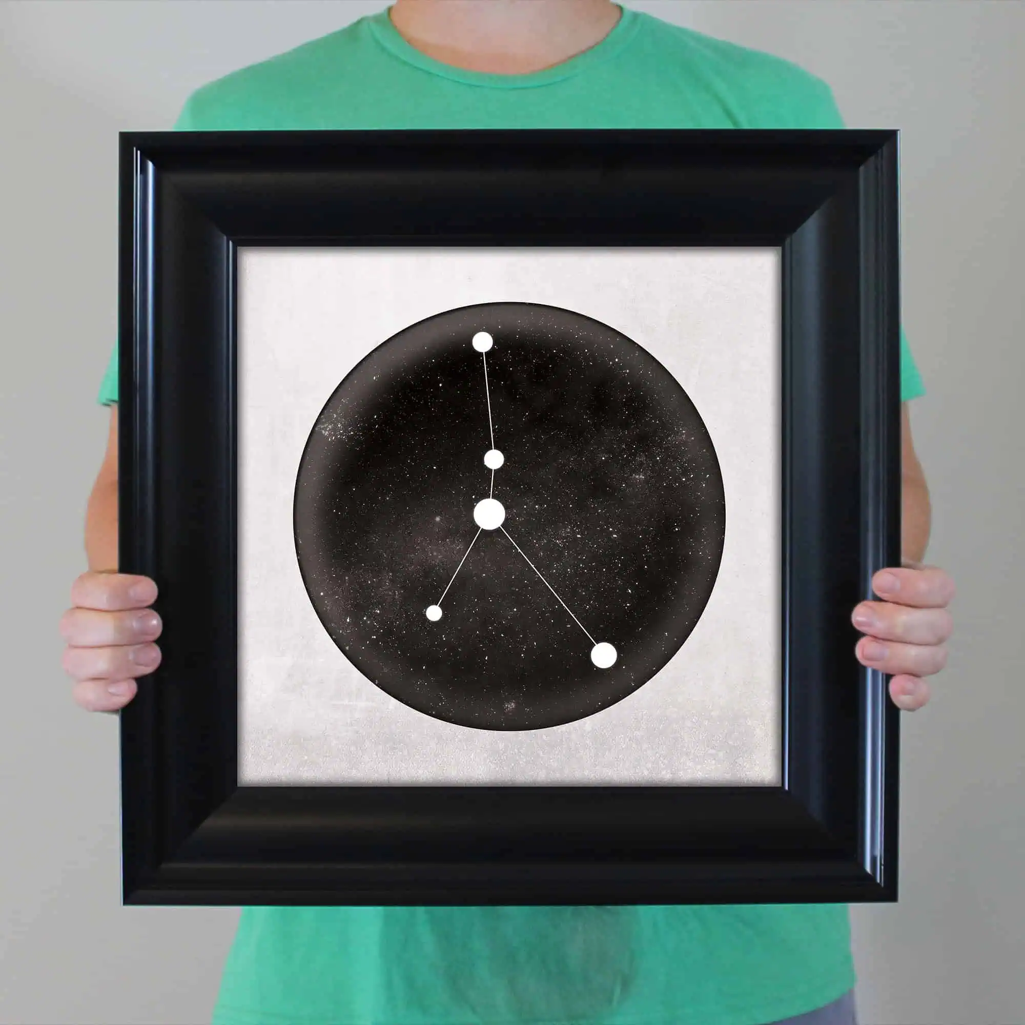 Constellation star map artwork featuring a black and white galaxy background, modern white dot and line design, framed and held by person in a green shirt.